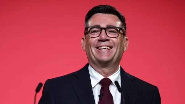 Andy Burnham souriant, fond rouge.