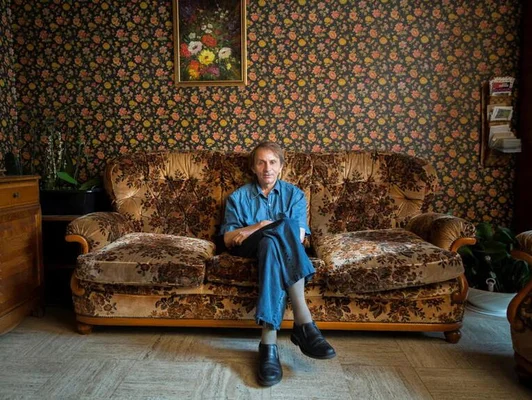 Portrait de Michel Houellebecq assis sur un canapé fleuri, vêtu d'une tenue en jean bleu.