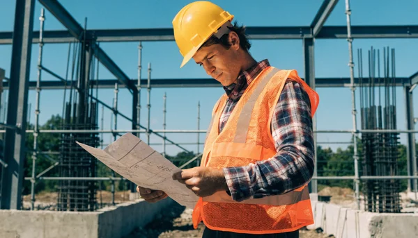 Homme d'une trentaine d'années, casque de chantier sur la tête, tenant un plan de construction sur un chantier ensoleillé. Plan rapproché, intention éditoriale : illustrer les métiers en pénurie ciblés par la prime trampoline, avec un ton pragmatique et constructif.