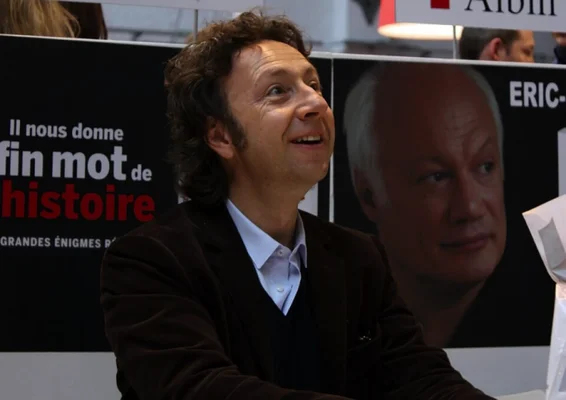 Stéphane Bern assis lors d'un événement, devant un panneau présentant un portrait et du texte.