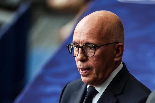 Éric Ciotti en costume sombre, devant un fond bleu.