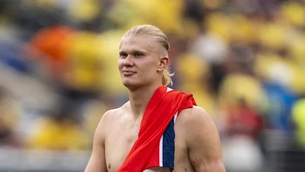 Erling Haaland, torse nu, drapé d'un drapeau, devant un stade en arrière-plan.