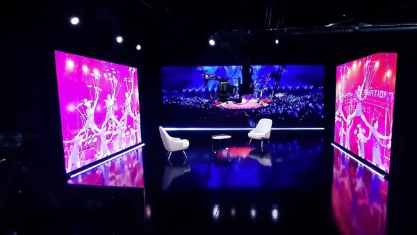 Plateau de tournage télé avec fauteuils blancs, table basse et écrans géants diffusant des images de spectacle.