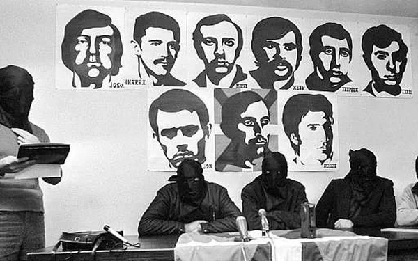 Membres d'ETA cagoulés lors d'une conférence de presse, avec des portraits d'autres membres en arrière-plan.