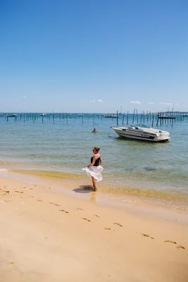 Femme contemplant la mer depuis la plage du Cap Ferret, bateaux et piquets ostréicoles visibles.