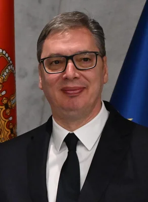 Le président serbe Aleksandar Vučić en costume devant un drapeau national et un autre drapeau.