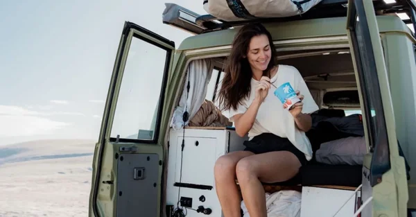 Femme souriant avec une pochette bleue et blanche dans l'encadrement d'un camping-car, paysage aride.