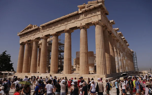 Le Parthénon sur l'Acropole d'Athènes sous un ciel bleu, avec des échafaudages et une foule de touristes au premier plan.