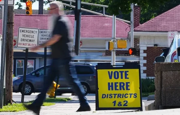 Un homme barbu passe devant un panneau 'VOTE HERE' sur un trottoir, évoquant le contexte électoral.
