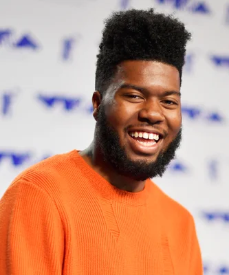 Khalid en pull orange, souriant devant un fond blanc.