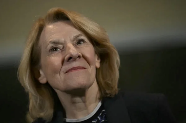 Portrait d'une femme âgée aux cheveux blonds, en blazer sombre, regardant au loin.