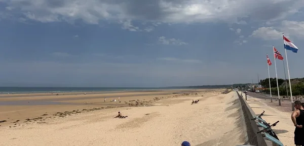 Plage du Débarquement par une journée ensoleillée, avec les drapeaux danois, norvégien et néerlandais flottant sur la promenade.