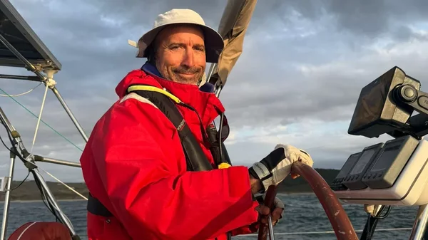 Julien Poublan à la barre de son voilier, souriant sous un ciel nuageux.