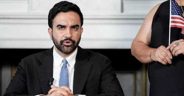 Zohran Mamdani s'adressant à la presse, avec un drapeau américain à ses côtés.