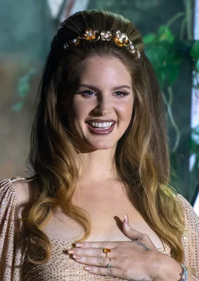 Lana Del Rey souriante, parée d'un bandeau floral doré et d'une robe pailletée.