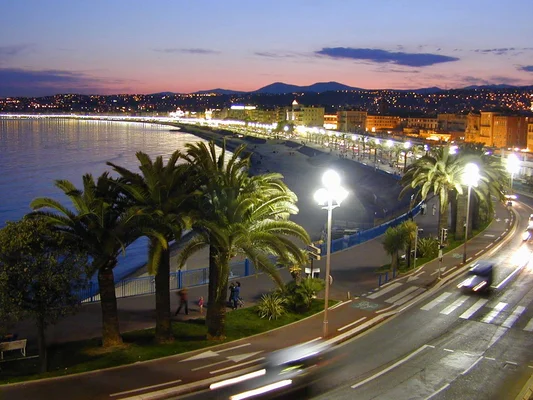 Vue nocturne de la Promenade des Anglais à Nice, illuminée par les lampadaires et les phares des voitures.