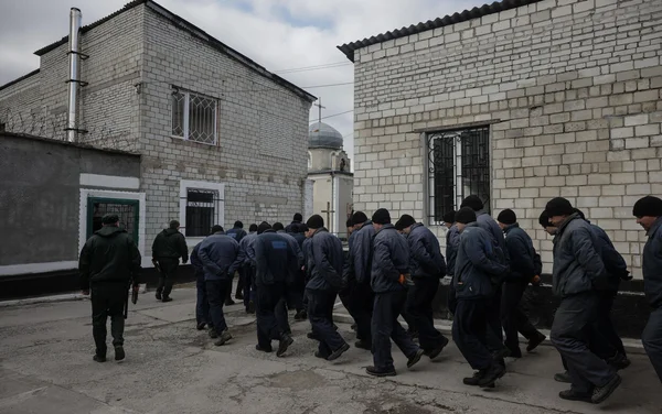Prisonniers russes en tenue bleue escortés dans la cour d'un camp pénitentiaire, avec une église en arrière-plan.