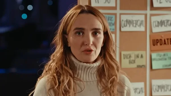 Chloe Fineman en pull blanc devant un tableau d'affichage.