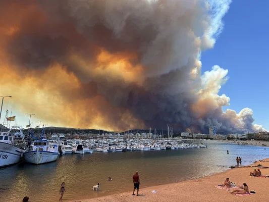Un immense panache de fumée noire provenant d'un incendie de forêt plane au-dessus d'une marina côtière et d'une plage où des gens se baignent.
