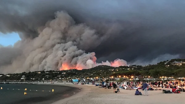 Un incendie de forêt ravage une colline surplombant une plage bondée au crépuscule, des spectateurs observent depuis le sable.
