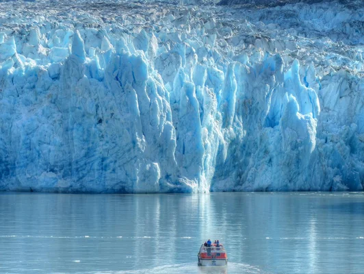 Un bateau orange avec des passagers devant une imposante paroi glaciaire en Antarctique, illustrant la fonte des glaces.