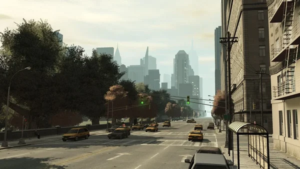 Une capture d'écran du jeu vidéo Grand Theft Auto IV (2008) montrant une vue à la troisième personne d'une rue de Liberty City. L'image présente un trafic routier dense avec plusieurs taxis jaunes, des immeubles résidentiels sur la droite et une ligne d'arbres sur la gauche. En arrière-plan, on distingue la silhouette brumeuse des gratte-ciels de la ville. La scène est baignée dans une lumière diurne caractéristique de l'ambiance visuelle du jeu.
