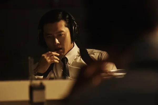 Plan moyen d'un homme asiatique, l'acteur Hyun Bin, portant un casque audio et une chemise blanche avec cravate. Il est assis dans un environnement sombre, éclairé par une lumière chaude et directionnelle, suggérant une scène de surveillance ou d'interrogatoire. Son expression est concentrée et sérieuse.