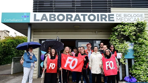 Groupe de grévistes portant des gilets rouges avec le logo FO posant devant l'entrée du laboratoire Cerballiance à Carcassonne, certains tenant des parapluies et un document.