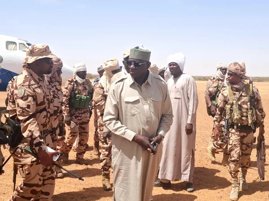 Groupe d'hommes, dont des militaires en tenue de camouflage et des civils en robes traditionnelles, rassemblés sur un aérodrome en terre près d'un avion blanc, au Tchad, après les affrontements intercommunautaires.