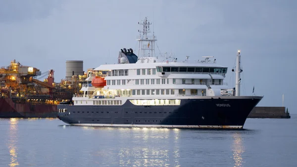Le navire d'expédition 'Hondius' naviguant dans un port au crépuscule, avec des structures industrielles illuminées en arrière-plan, représentant le navire touché par le hantavirus aux Canaries.