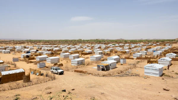 Vue d'ensemble d'un camp de tentes blanches et d'abris de fortune dans le paysage aride de l'est du Tchad, avec un SUV blanc garé au milieu, illustrant les conditions de vie des communautés touchées par l'affrontement intercommunautaire.