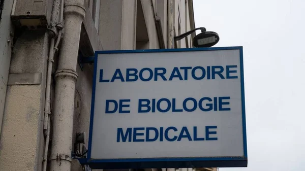 Panneau blanc avec inscription bleue 'LABORATOIRE DE BIOLOGIE MEDICALE' fixé sur un mur extérieur, à côté de tuyaux verticaux et sous un lampadaire.