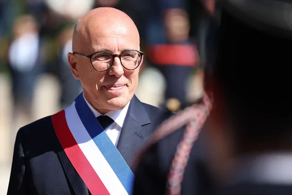 Éric Ciotti, arborant l'écharpe tricolore, souriant en costume sombre, regardant vers la droite devant un fond flou