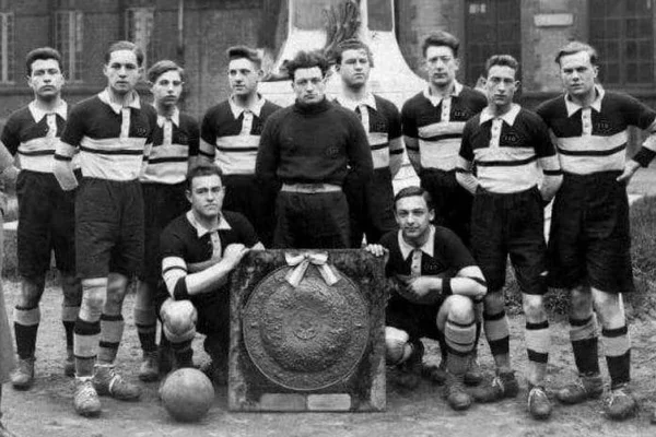 Photographie en noir et blanc d'une équipe de football posant avec un grand trophée bouclier, joueurs en maillots rayés et gardien en pull sombre.