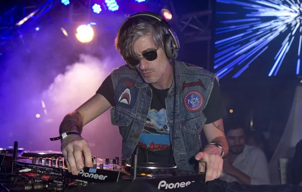 Kavinsky, DJ aux cheveux grisonnants et lunettes de soleil, en gilet denim à patchs, mixant sur des platines Pioneer sous un éclairage bleu de scène