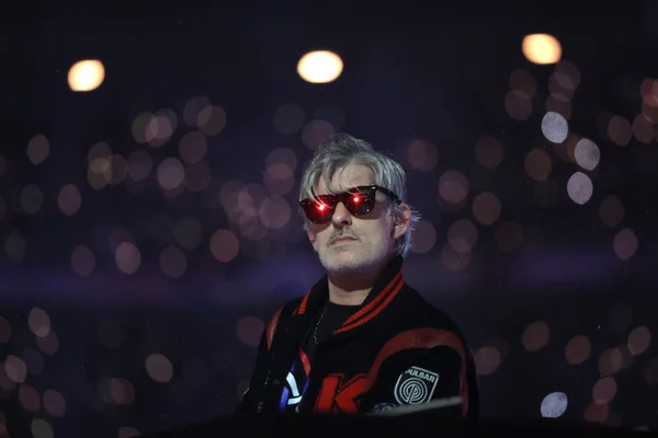 Kavinsky, le DJ français de « Nightcall », vu en plan moyen avec des cheveux gris et des lunettes de soleil reflétant des lumières rouges, sur un fond sombre avec des effets de bokeh