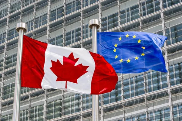 Les drapeaux du Canada et de l'Union européenne flottant côte à côte sur des mâts argentés devant un immeuble moderne en verre à façade en grille, symbolisant la coopération transatlantique.