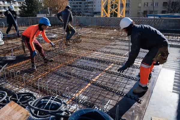 Groupe d'ouvriers du bâtiment portant casques et équipements de sécurité sur un toit, disposant des grilles de ferraillage pour une coulée de béton, avec une grue visible en arrière-plan.