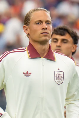 Marcos Llorente, joueur de football espagnol aux cheveux blonds longs attachés, vêtu d'un survêtement blanc et bordeaux de l'équipe nationale d'Espagne, regarde droit devant lui avec une expression concentrée, flou d'un autre joueur en arrière-plan.