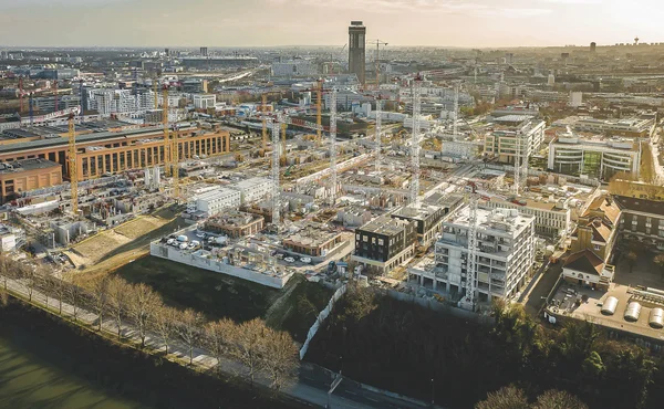 Vue aérienne du chantier de construction du village des athlètes pour les JO 2024, avec de nombreuses grues et des bâtiments en cours d'édification, en arrière-plan d'une ville existante sous un ciel brumeux.