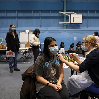 Une jeune femme portant un t-shirt gris et un masque facial reçoit un vaccin dans le bras d'une professionnelle de santé dans un gymnase, tandis que d'autres personnes masquées attendent en arrière-plan, dans le cadre de la lutte contre une épidémie de méningite en Angleterre.