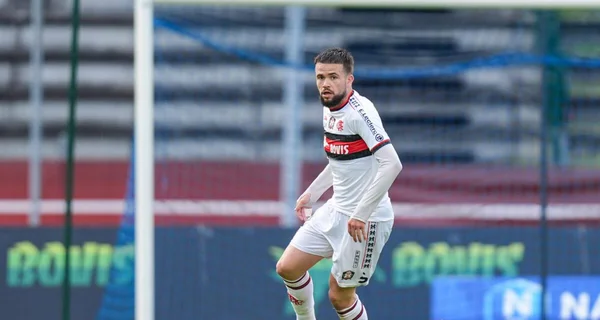 Un joueur de football barbu en maillot blanc à bandes rouges et noires courant sur le terrain lors d'un match de Ligue 3, avec des tribunes et des panneaux publicitaires flous en arrière-plan