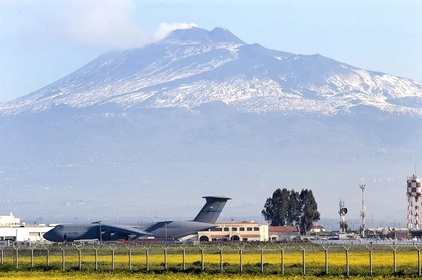 Un avion de transport militaire américain C-130 stationné sur le tarmac de la base aérienne de Sigonella, en Sicile, avec l'Etna enneigé en arrière-plan sous un ciel brumeux.