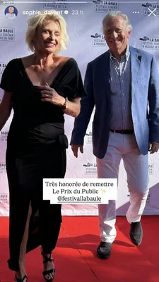Sophie Davant en robe portefeuille noire fendue et talons aiguilles au bras de William Leymergie sur le tapis rouge du Festival de La Baule, avec un encart texte blanc sur leurs jambes.