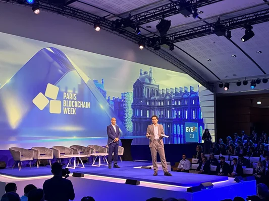 Deux intervenants en costume sur la scène de la Paris Blockchain Week, devant un grand écran affichant le logo de l'événement et un décor d'architecture parisienne, face à un public assis dans la salle.