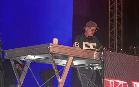Kavinsky, casquette et lunettes de soleil, en sweat noir à lettrage blanc, derrière son setup DJ sur scène avec un éclairage bleu en arrière-plan