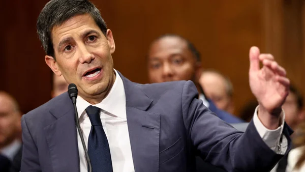 Kevin Warsh en costume sombre et cravate, s'exprimant devant un micro avec la main droite levée, dans une salle d'audience du Sénat américain, entouré de personnes assises.