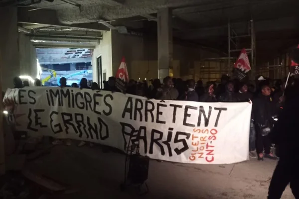 Manifestation nocturne sur un chantier de l'Arena pour les JO 2024 : un groupe de protestataires tient une banderole blanche avec le texte 'LES IMMIGRÉS ARRÊTENT LE GRAND PARIS' dans un bâtiment en béton non fini, éclairé faiblement.