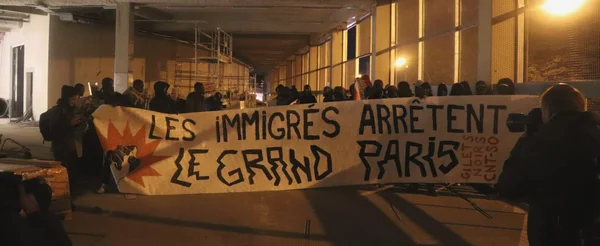 Grève des travailleurs immigrés sur les chantiers des JO : un groupe de manifestants tient une grande banderole 'LES IMMIGRÉS ARRÊTENT LE GRAND PARIS' dans un passage souterrain urbain, avec des échafaudages en arrière-plan.