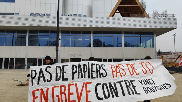 Manifestants soutenant les travailleurs sans-papiers devant un bâtiment moderne 'GYMNASES' sur un chantier des JO 2024, tenant une banderole 'PAS DE PAPIERS PAS DE JO' et 'EN GREVE CONTRE VINCI BOUYGUES'.